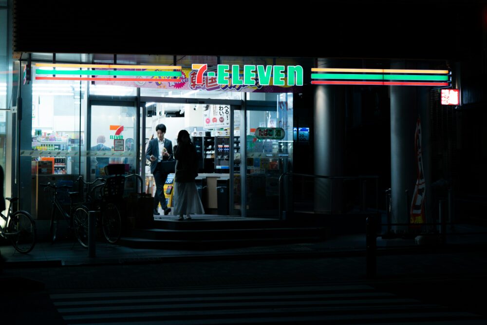 夜の街で明るく光るコンビニの入り口と、そこへ吸い寄せられるように歩く人々のイメージ